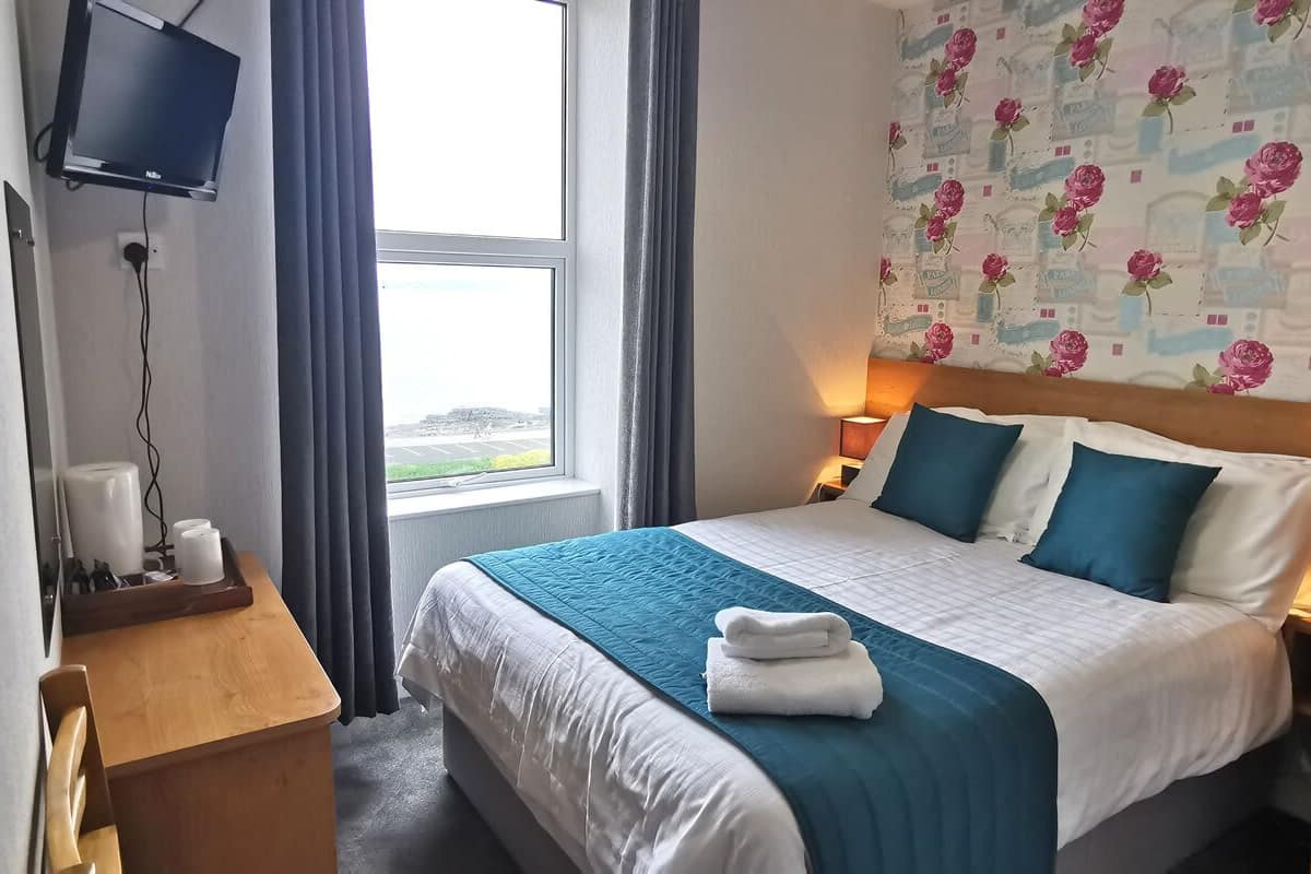 A hotel room with a bed, a TV mounted on the wall, a window with curtains, and a floral wallpaper.