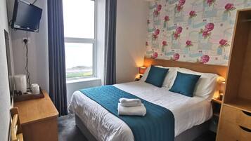Ein Hotelzimmer mit Bett, Wandfernseher, Fenster mit Vorhängen und blumigem Tapetenmuster.