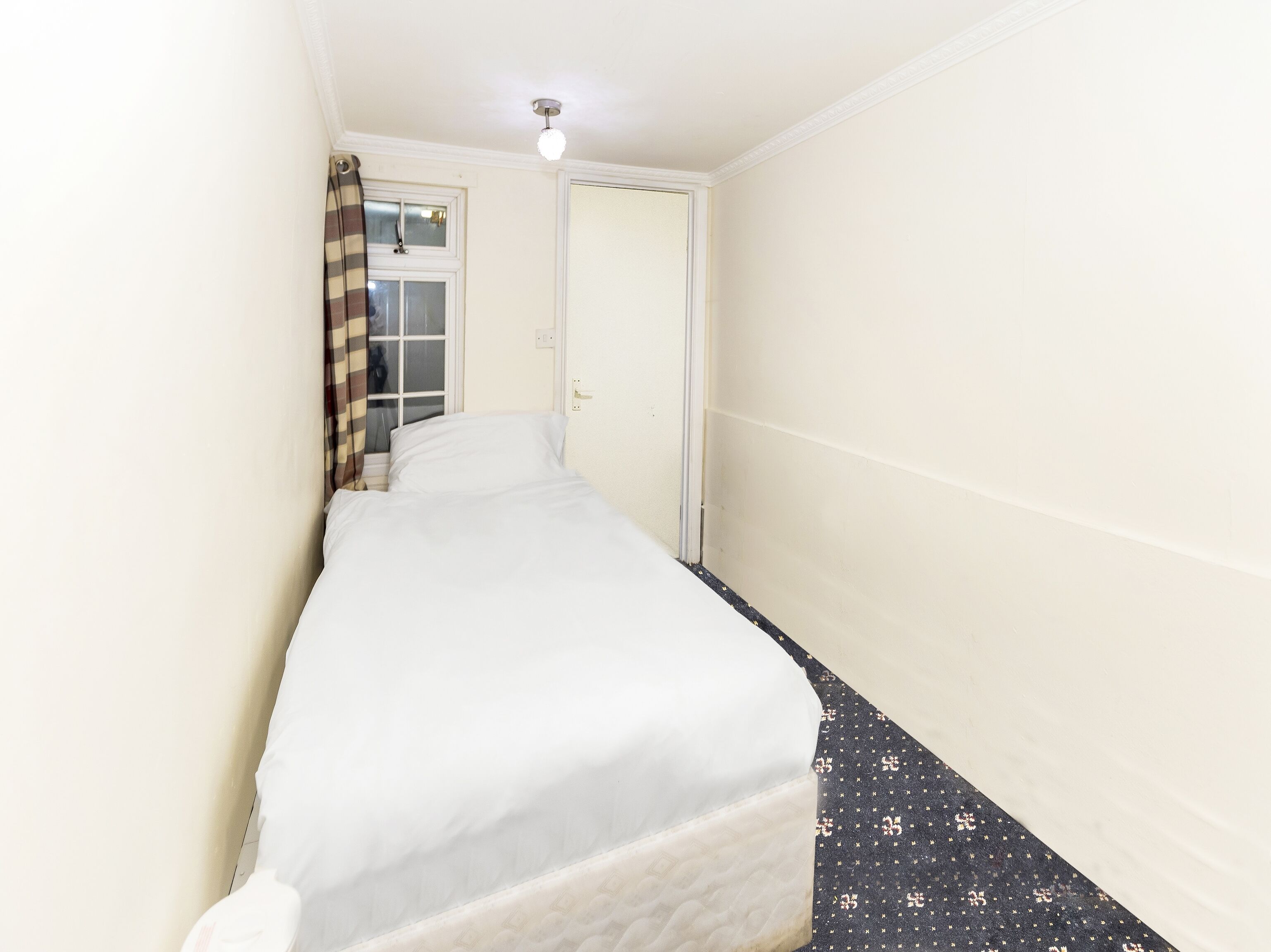 A narrow hotel room with a single bed, a window with curtains, and a door.