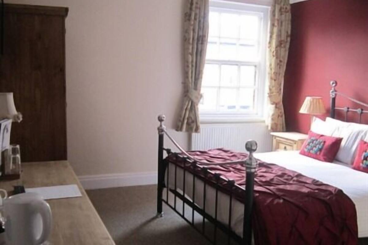 Quarto com cama, mesa de cabeceira, abajur e janela com cortinas.