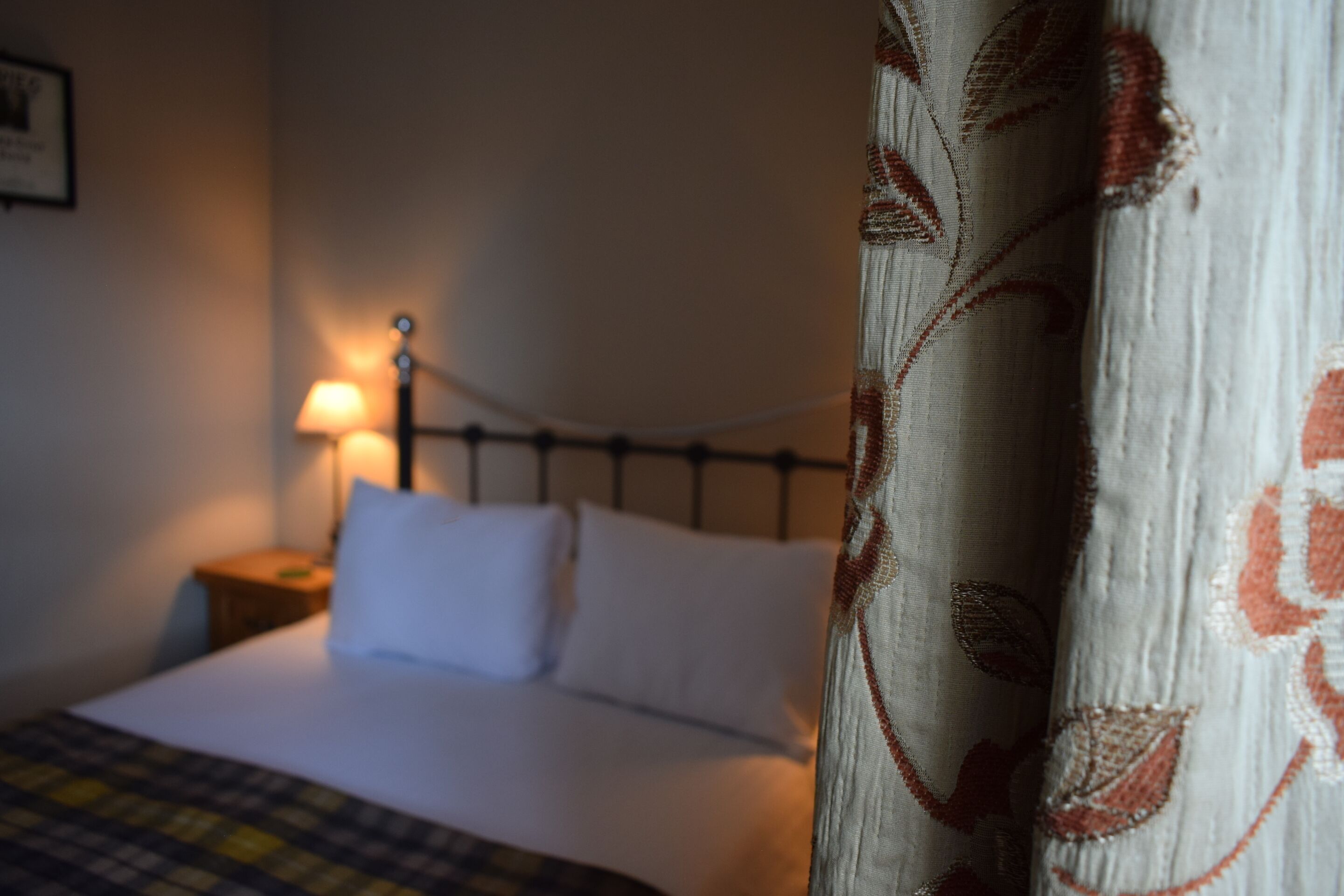 Ein Bett mit weißer Bettwäsche, ein Nachttisch mit Lampe und ein gemustertes Vorhang.