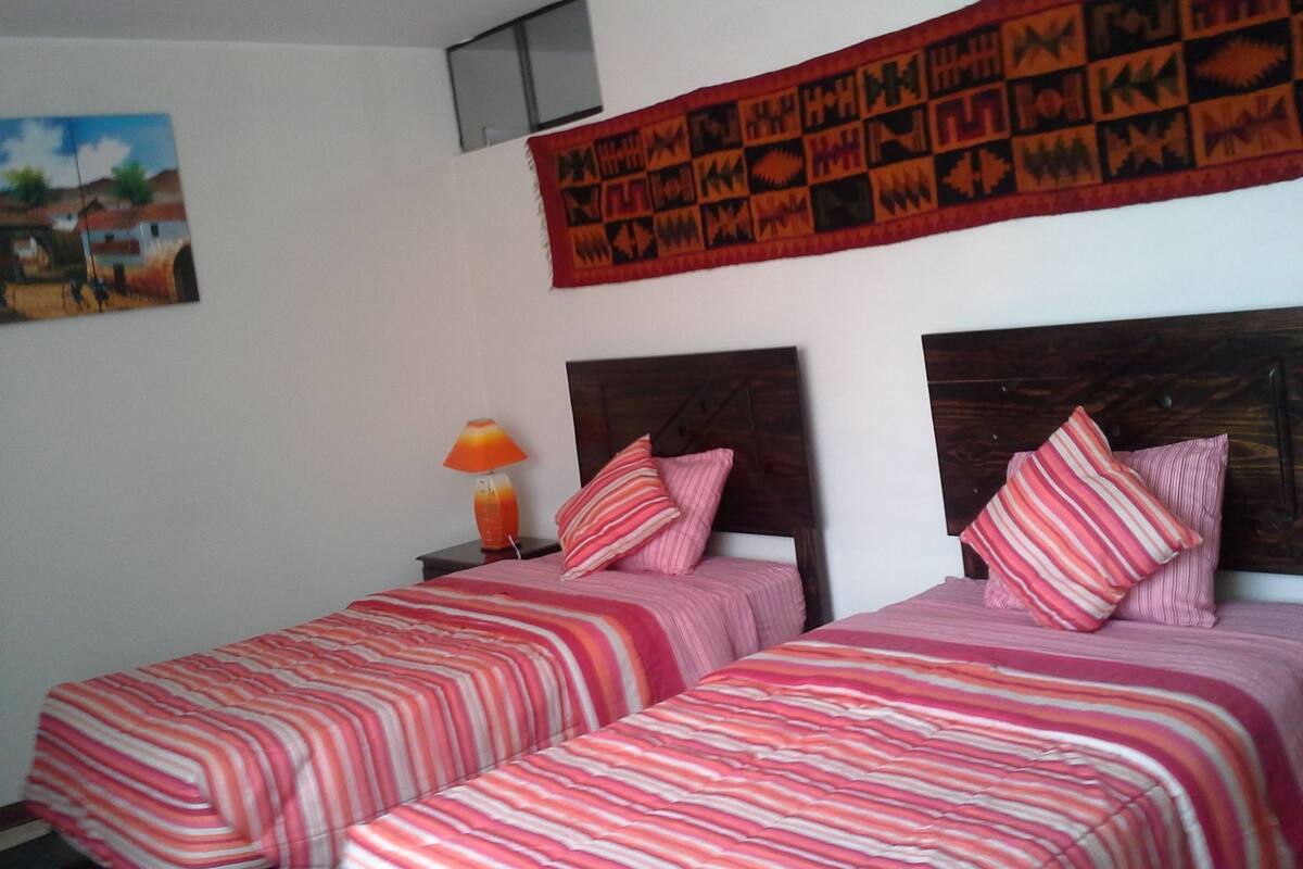 A bedroom with two beds, each with striped bedding, a nightstand with a lamp, and a tapestry on the wall.