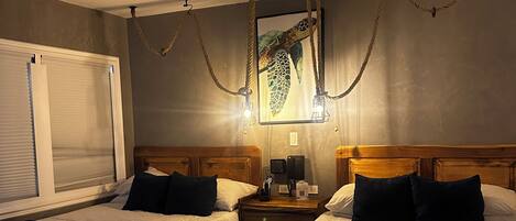 Habitación de hotel con dos camas, un ventanal grande con persianas, cabecera de madera, un banco tejido y un cuadro en la pared.