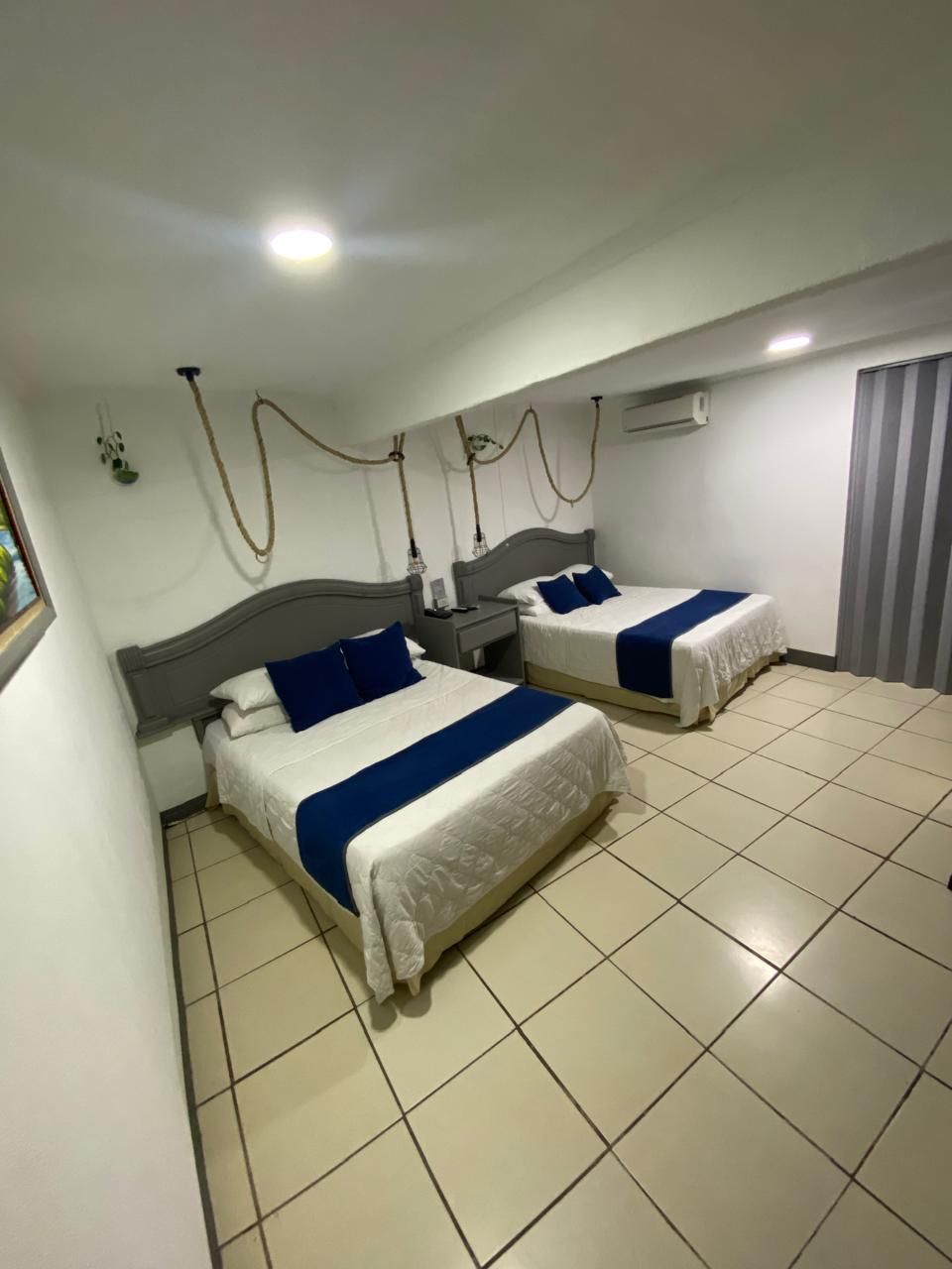 Ein Hotelzimmer mit zwei Betten, beide mit blauen Akzenten, weißer Bettwäsche und grauem Kopfteil. An jedem Bett befindet sich ein Nachttisch mit einer Lampe und ein kleiner Tisch. Der Raum hat einen Fliesenboden und ein Fenster mit Jalousien.