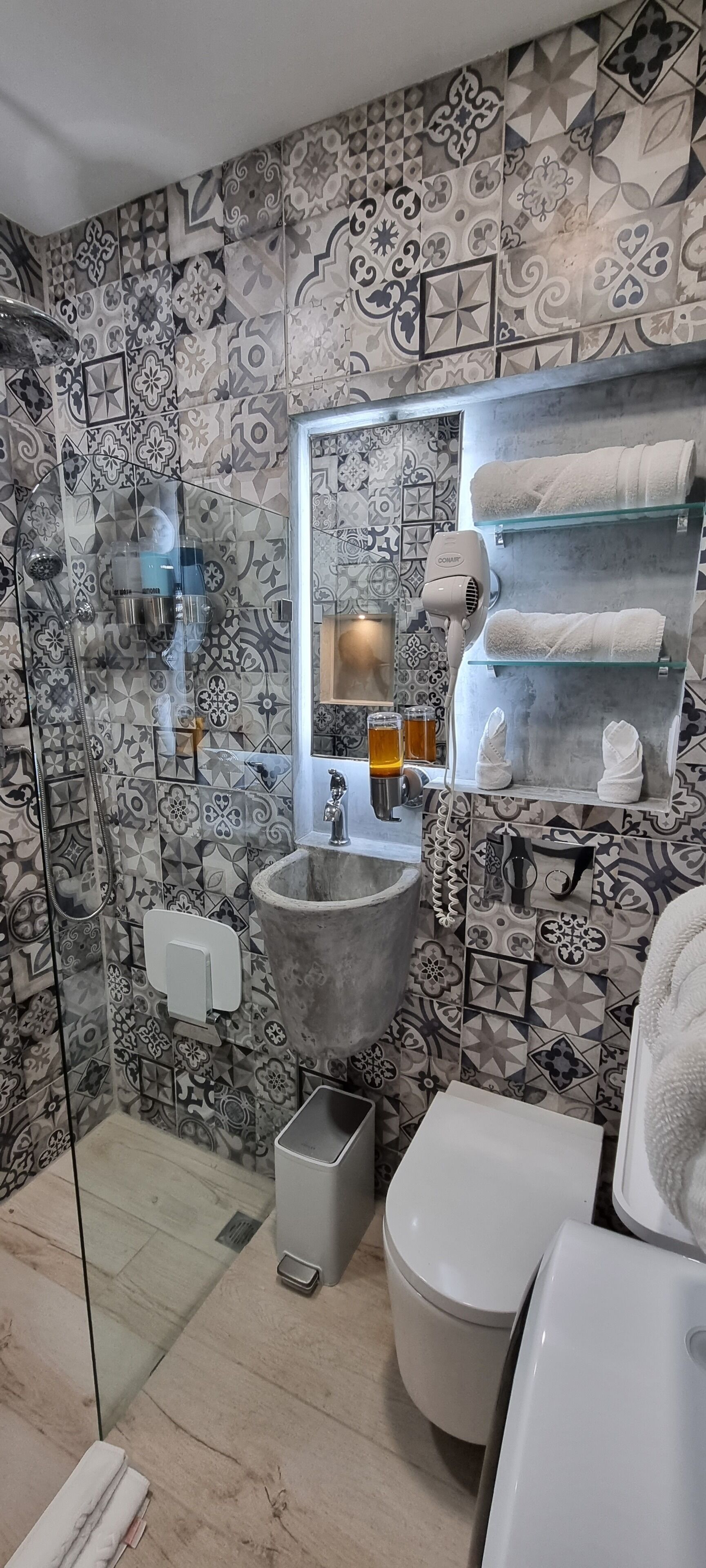 A bathroom with patterned tiles, a large sink, a glass shower enclosure, a toilet, and a towel rack with towels.