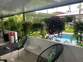 Un balcón con dos sillas de alambre negras, una mesita blanca y varios maceteros con plantas.