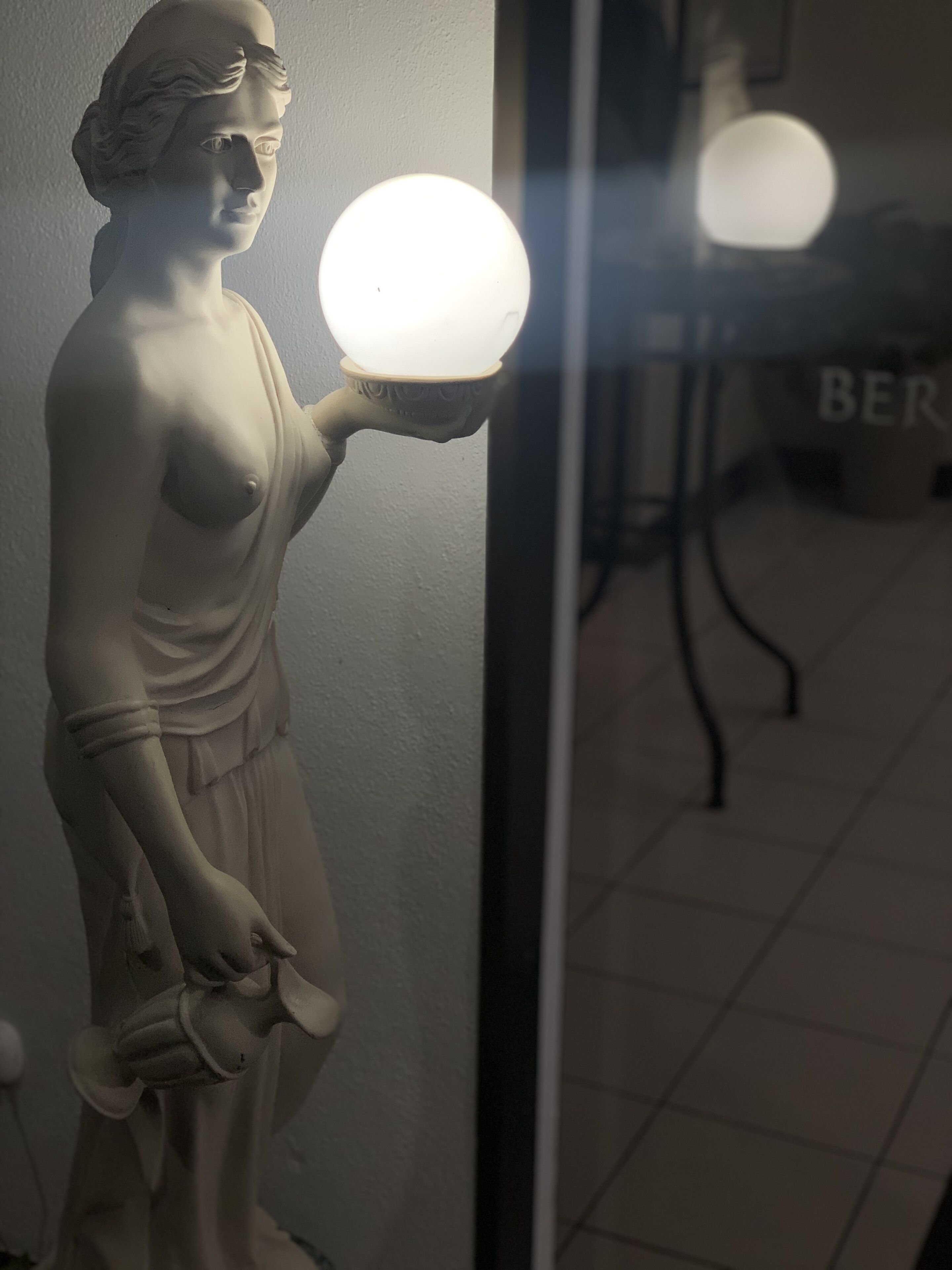 A statue of a woman holding a light bulb, positioned next to a mirror reflecting a chair and a wall with the word "BER".