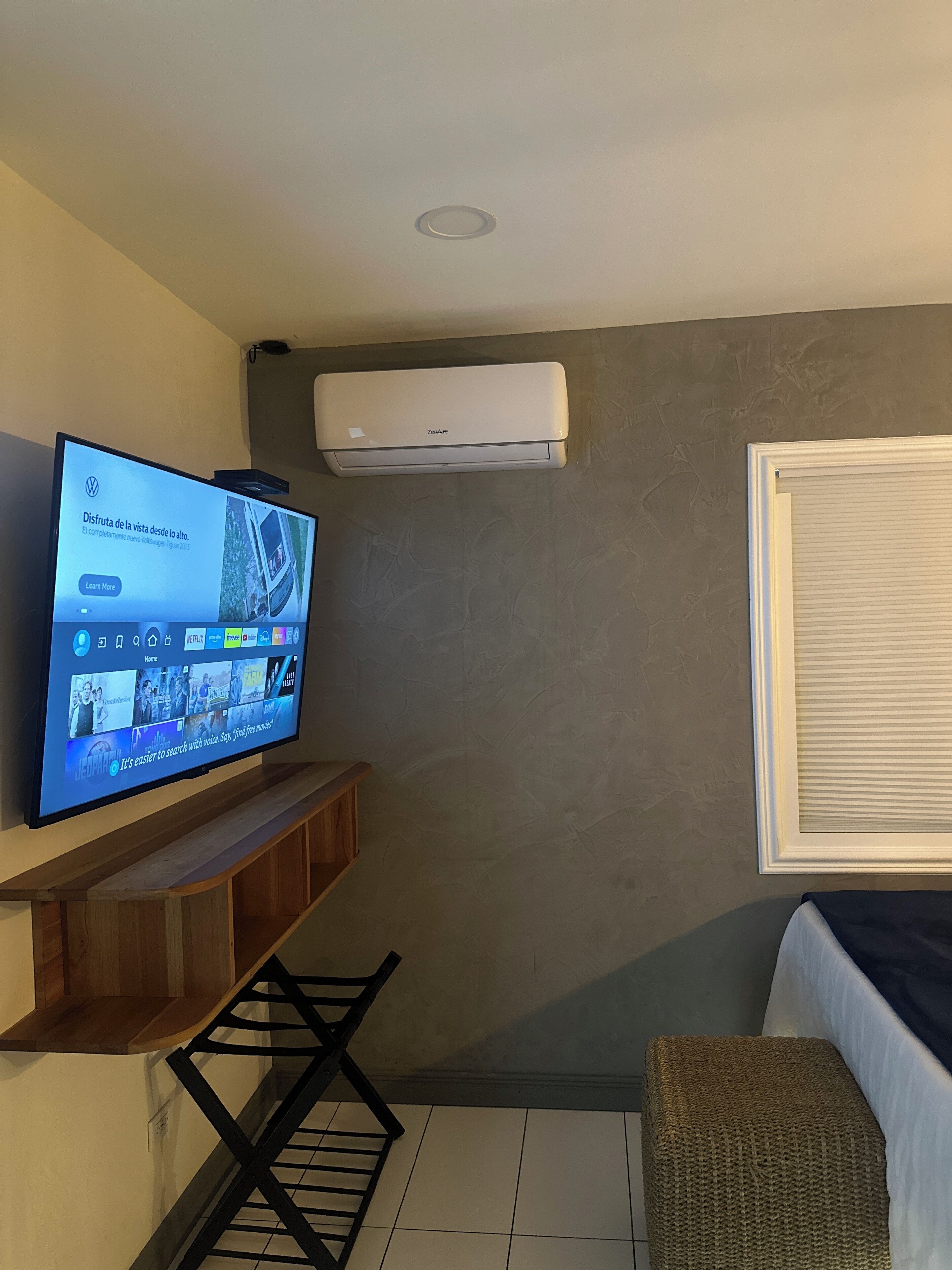 A living room with a flat-screen TV mounted on a wooden shelf, an air conditioning unit on the wall, a window with blinds, and a woven ottoman.