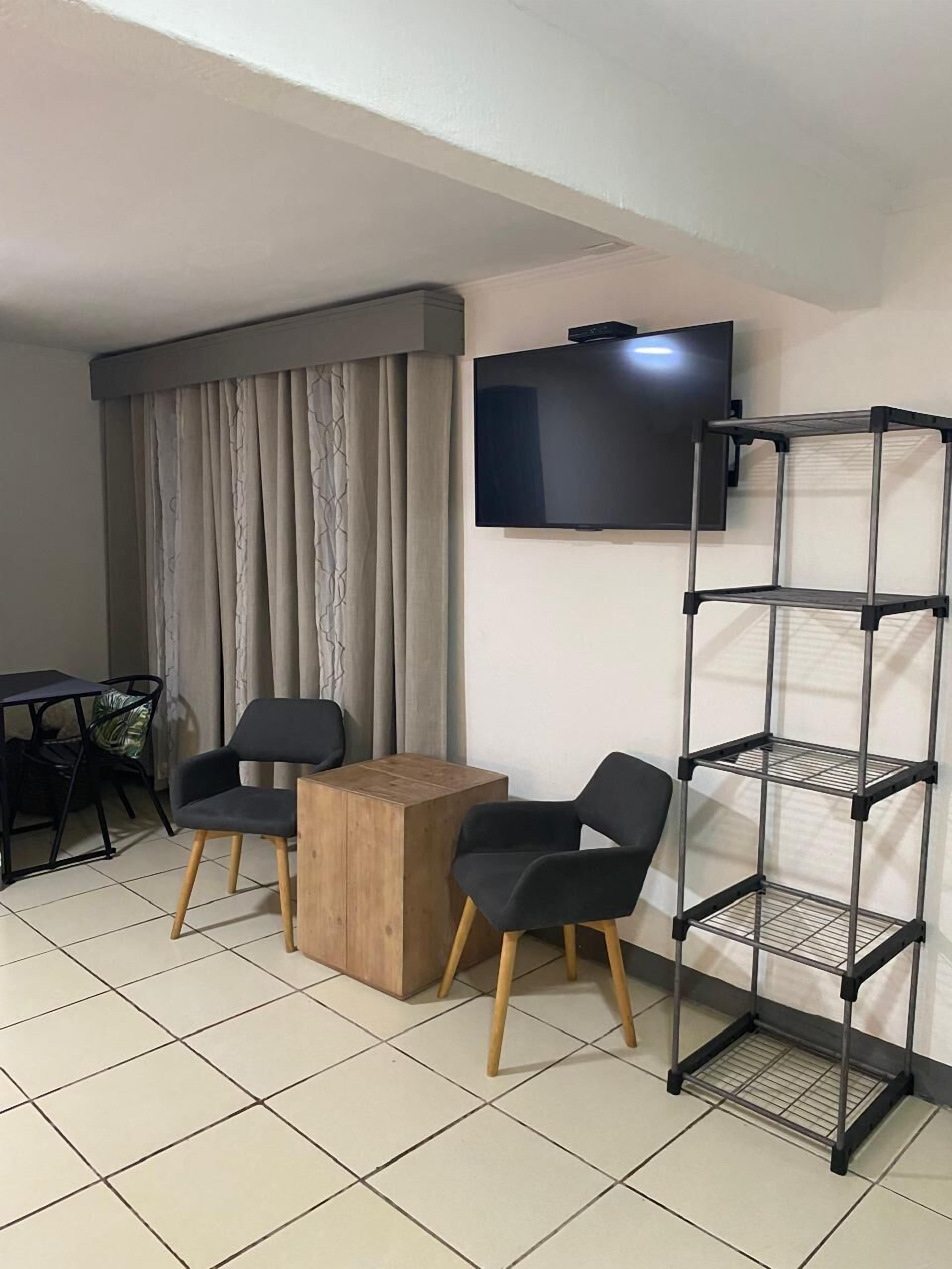 A room with a TV mounted on the wall, a wooden table, two chairs, a metal shelf unit, and a curtain.