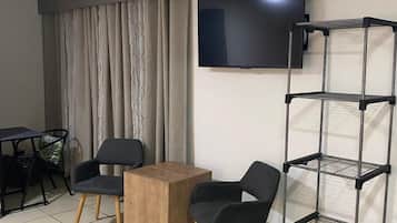 A room with a TV mounted on the wall, a wooden table, two chairs, a metal shelf unit, and a curtain.