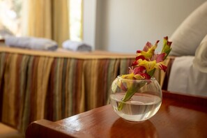 A clear glass vase with water and flowers on a wooden table in a room with striped curtains and a sofa.
