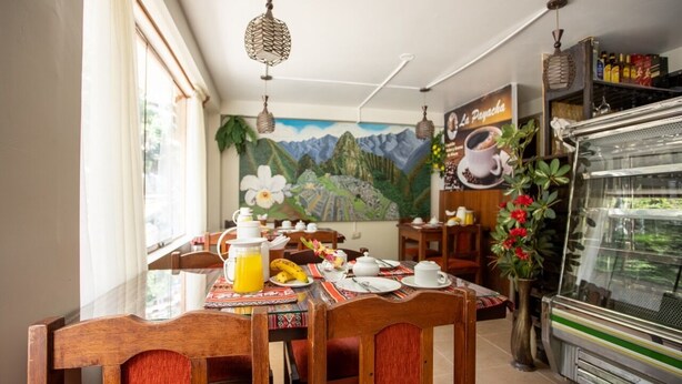 A dining area with wooden chairs and a table set with breakfast items, a mural of mountains, and a coffee machine. - Hostal La Payacha (Machu Picchu)