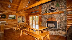 A wooden lodge interior with a stone fireplace, wooden benches, and a large dining table.