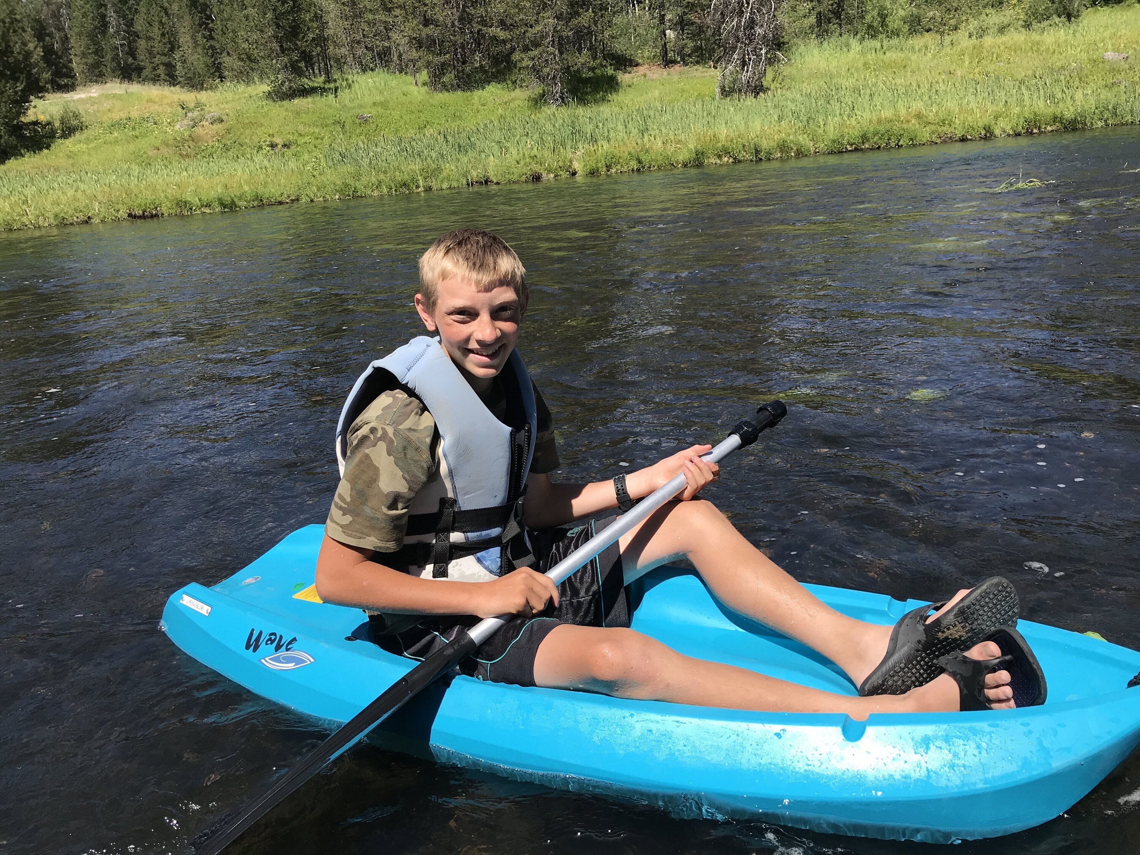 Une personne est assise sur un radeau gonflable bleu et tient une pagaie. Elle porte un gilet de sauvetage et des sandales, et se trouve sur une rivière bordée de verdure.