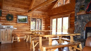 A log cabin interior with wooden furniture, stone fireplace, and large windows.