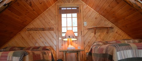 A cozy attic bedroom with two beds, a table with a lamp, and a window with blinds.