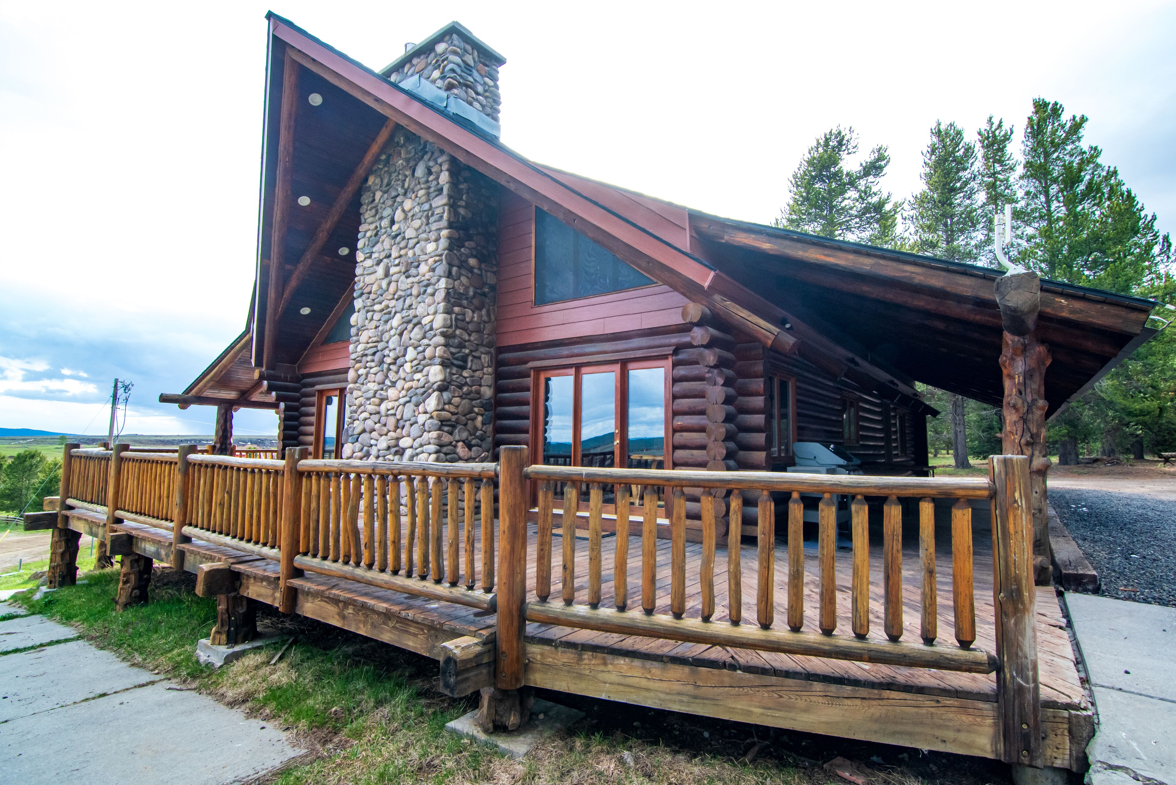 A log cabin with a stone chimney, wooden deck, and a large window overlooking a scenic landscape.