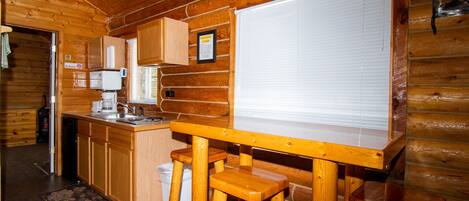 A wooden cabin interior with a kitchen area, wooden stools, a whiteboard, and a window with blinds.