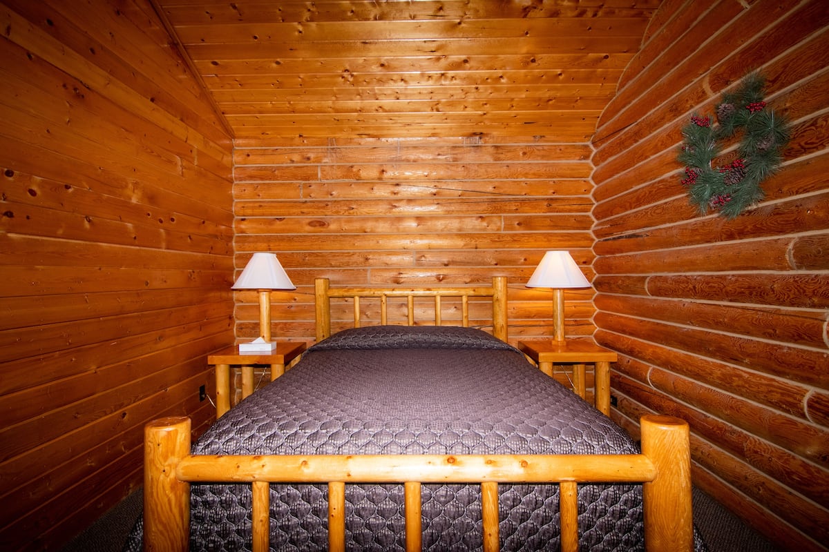 A wooden cabin room with a bed, two bedside lamps, and a Christmas wreath on the wall.