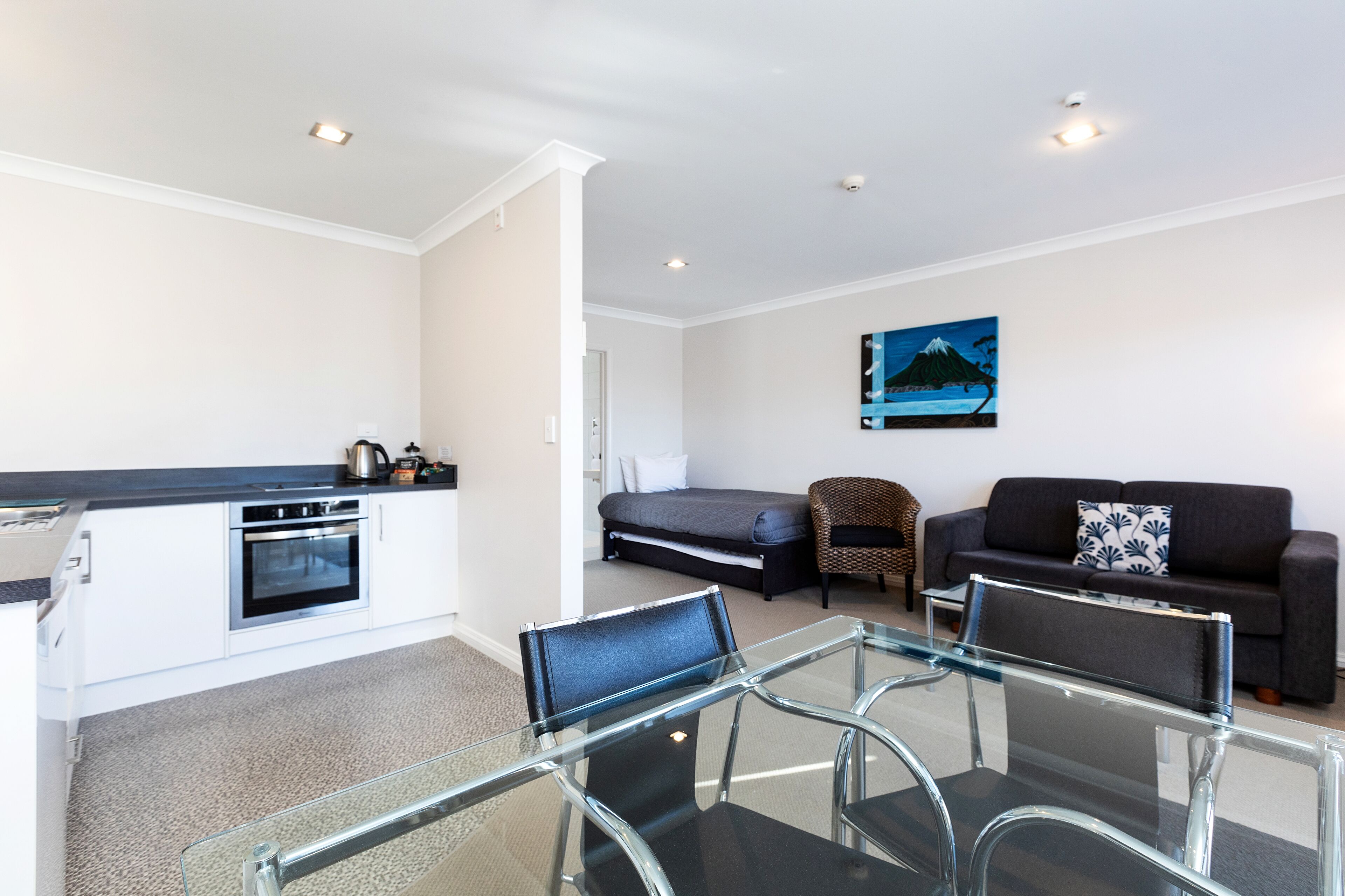 A modern kitchen with a built-in oven, a dining area with a glass table and chairs, a sofa, and a painting on the wall.