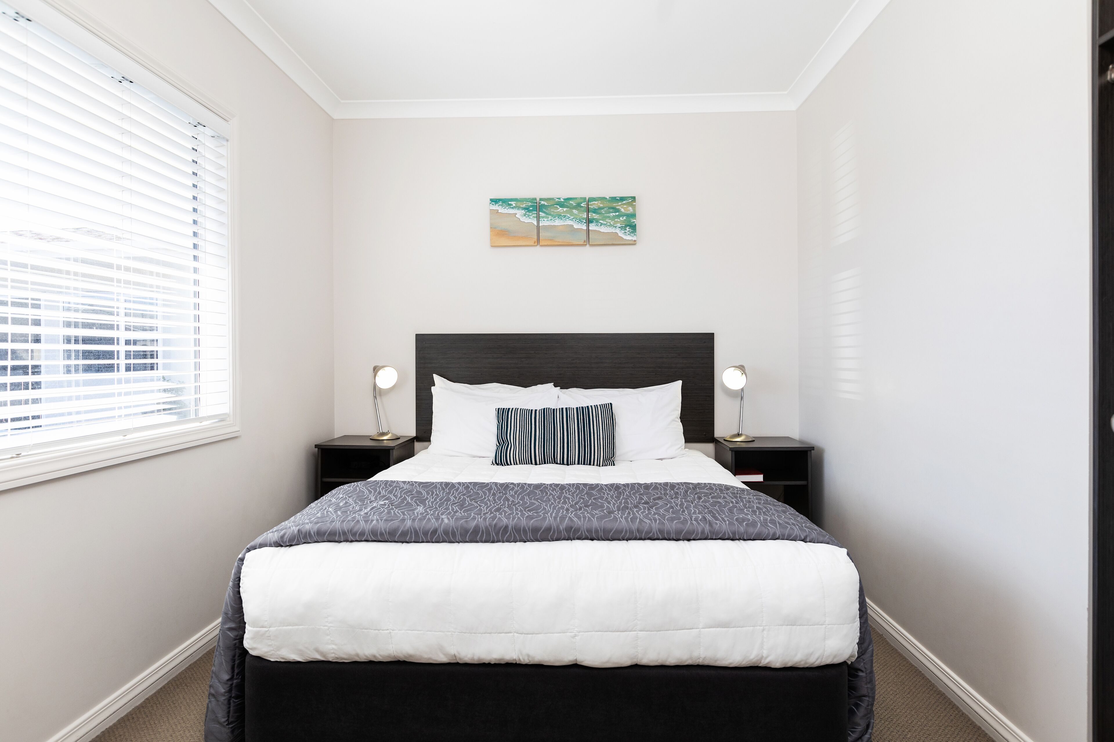 A neatly made bed with a white and gray comforter, a dark headboard, and a small bedside table with a lamp.