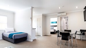 Une chambre d’hôtel moderne comprenant un lit, une table en verre et des chaises, une salle de bain avec douche et toilettes, et une grande fenêtre.