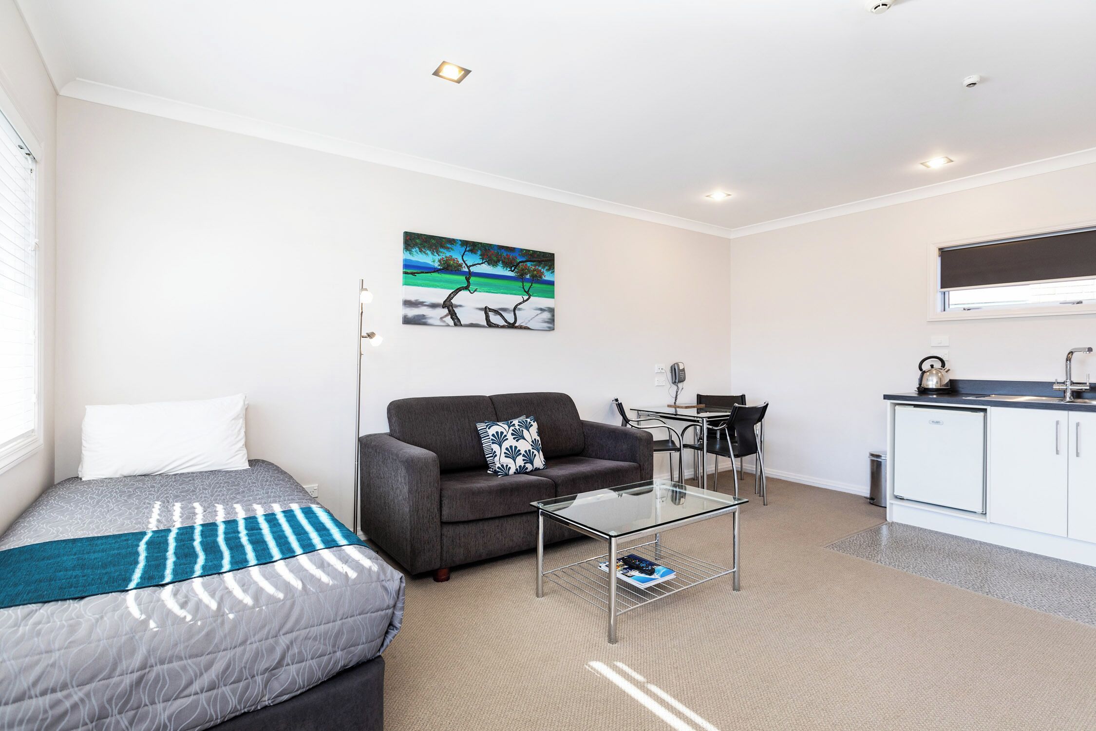 A modern room with a bed, sofa, glass coffee table, and a kitchenette area.