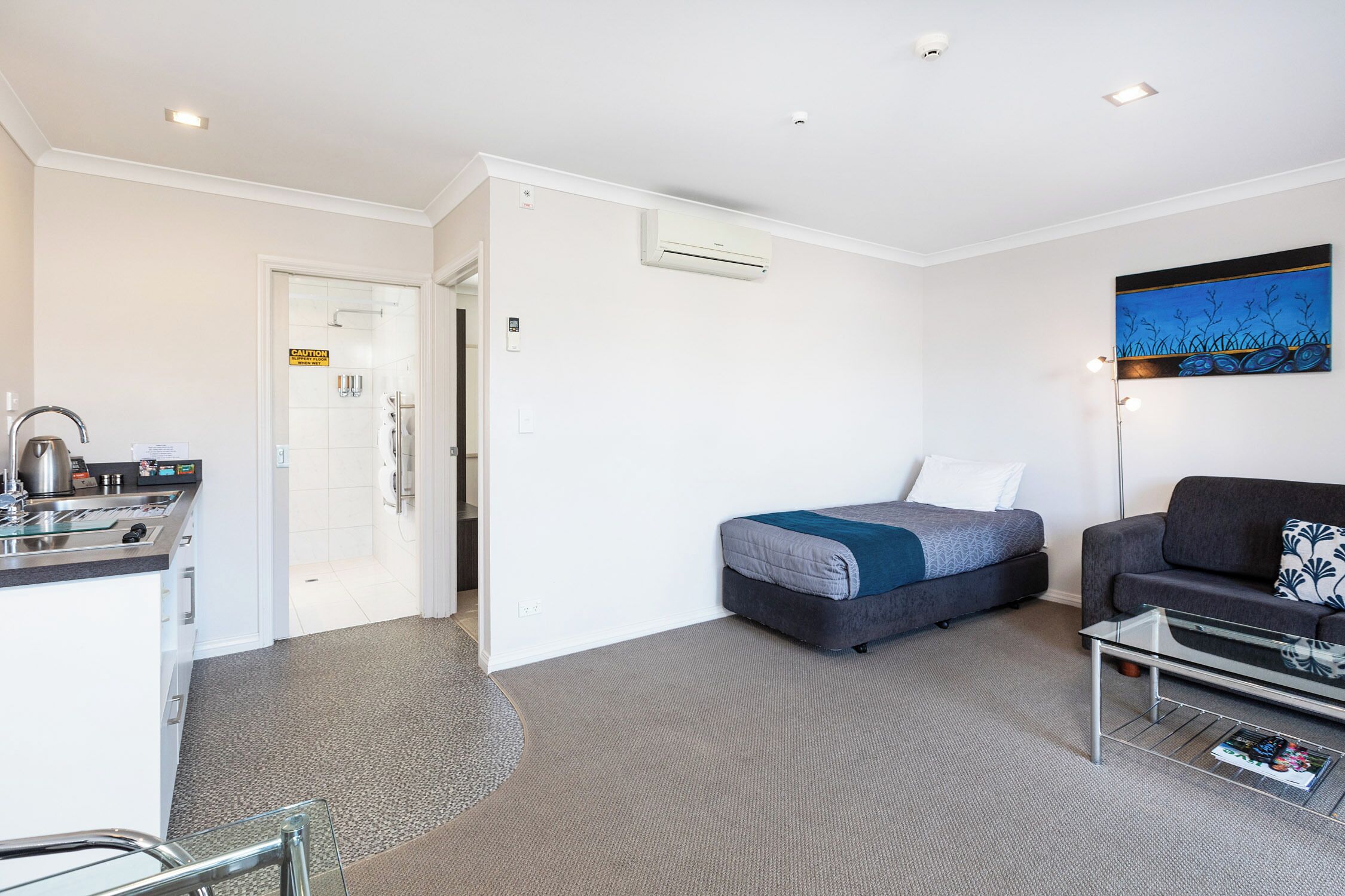A hotel room with a kitchenette, a bed with a blue blanket, a gray sofa, and a painting on the wall.