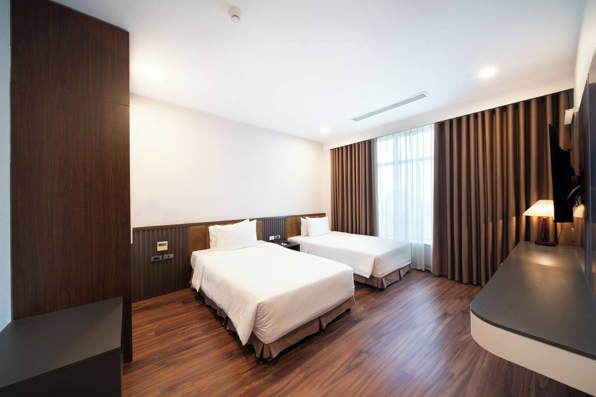 Habitación de hotel con dos camas, suelo de madera, televisor de pantalla plana y un ventanal con cortinas.