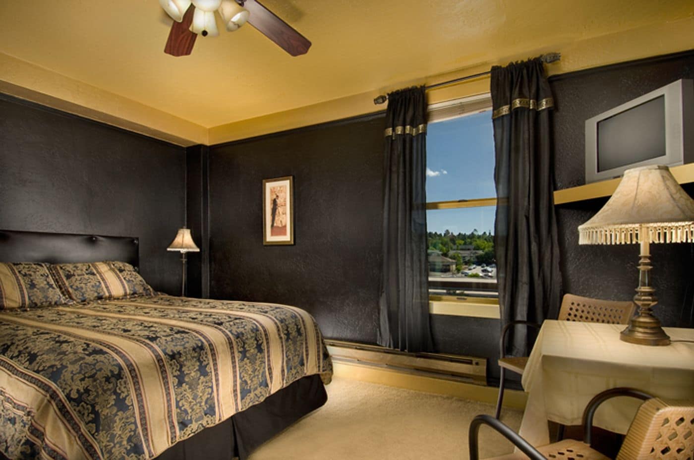 Une chambre d’hôtel avec un lit, une table avec une lampe, une chaise, une fenêtre avec des rideaux et un ventilateur de plafond.
