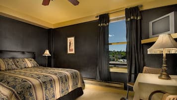 Une chambre d’hôtel avec un lit, une table avec une lampe, une chaise, une fenêtre avec des rideaux et un ventilateur de plafond.