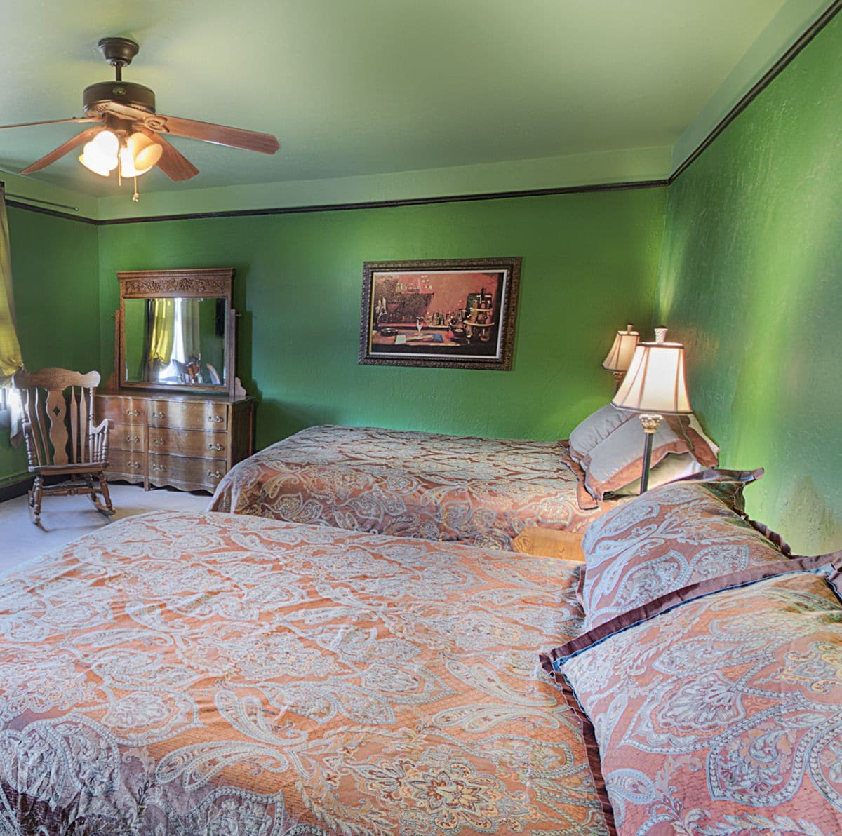 A room with two beds, a dresser, a chair, a ceiling fan, a framed picture, and a lamp.