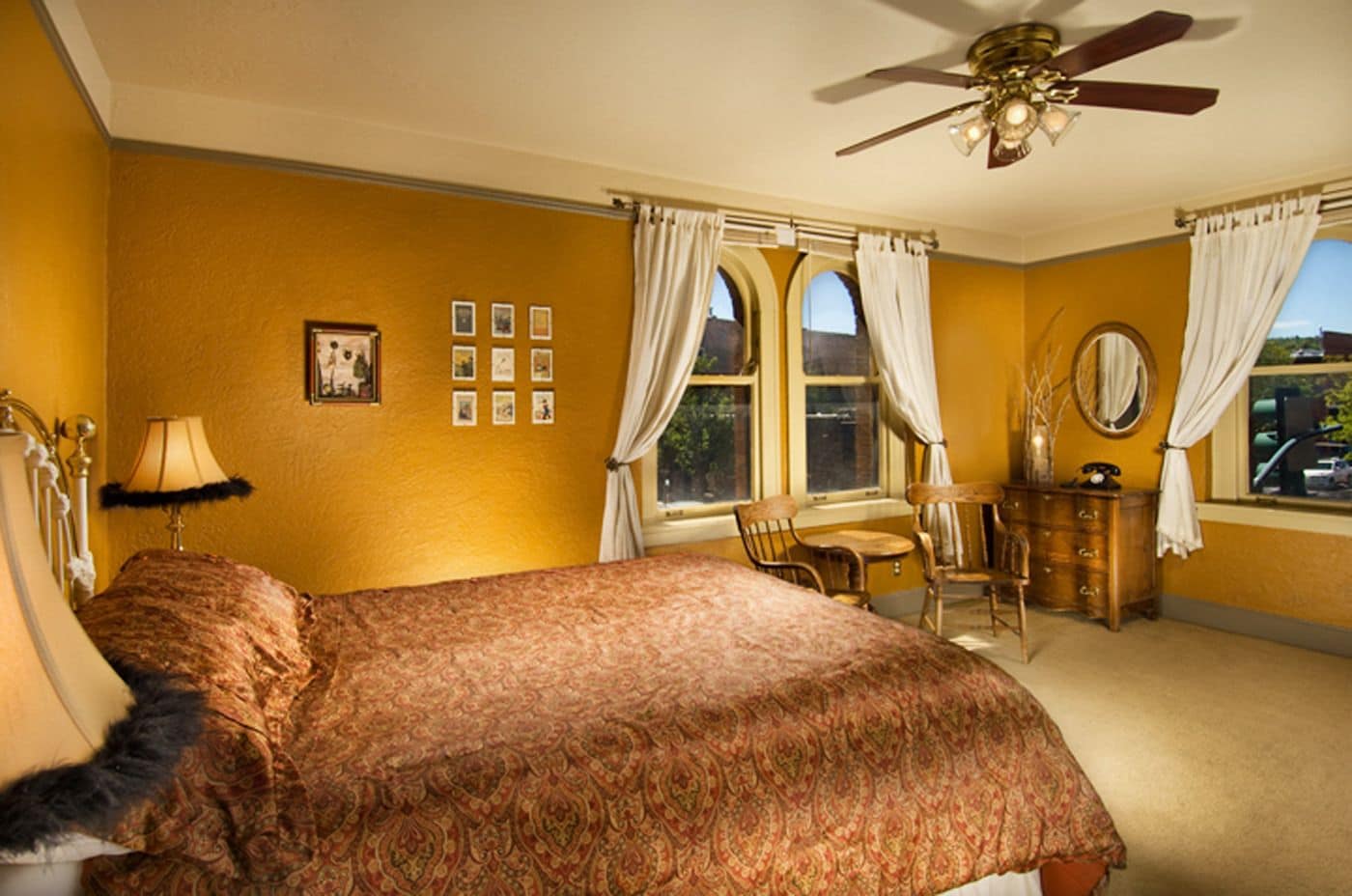 Une chambre à coucher avec un grand lit, un ventilateur de plafond, une commode, une chaise et une fenêtre avec des rideaux.