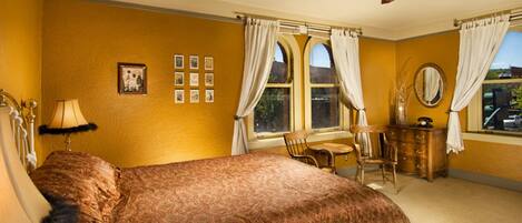 A bedroom with a large bed, a ceiling fan, a dresser, a chair, and a window with curtains.