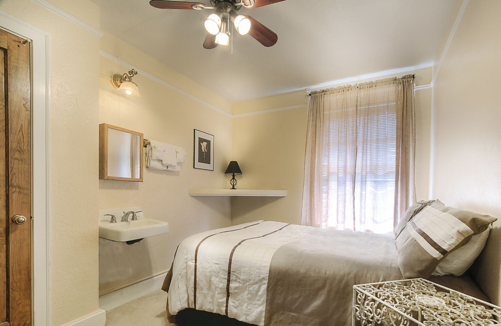 Une chambre à coucher comprenant un lit, une table de chevet, un ventilateur de plafond, une fenêtre avec des rideaux, une étagère fixée au mur et une porte.