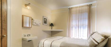 A bedroom with a bed, a bedside table, a ceiling fan, a window with curtains, a wall-mounted shelf, and a door.