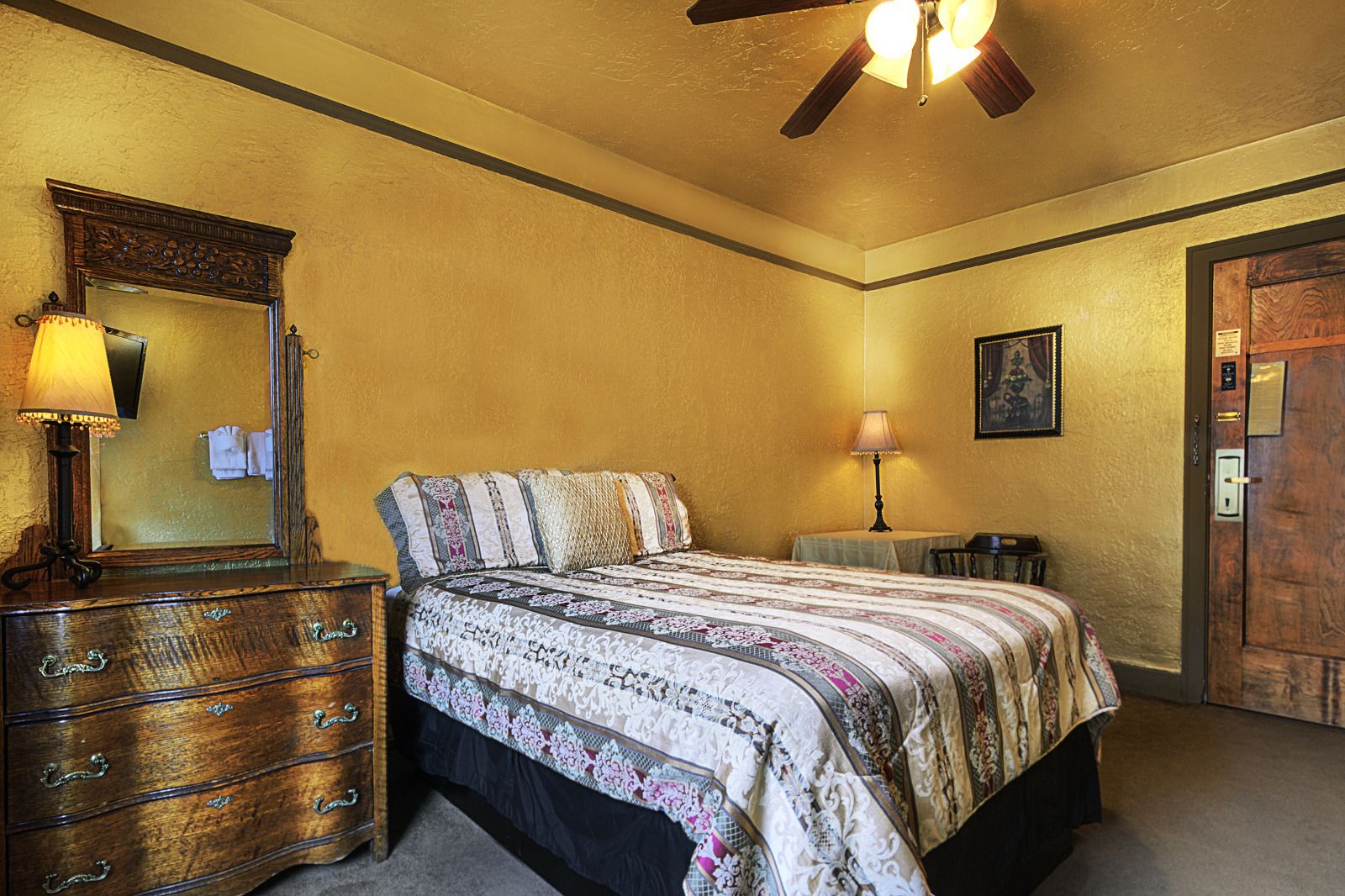 A bedroom with a bed, dresser, mirror, ceiling fan, and a door.