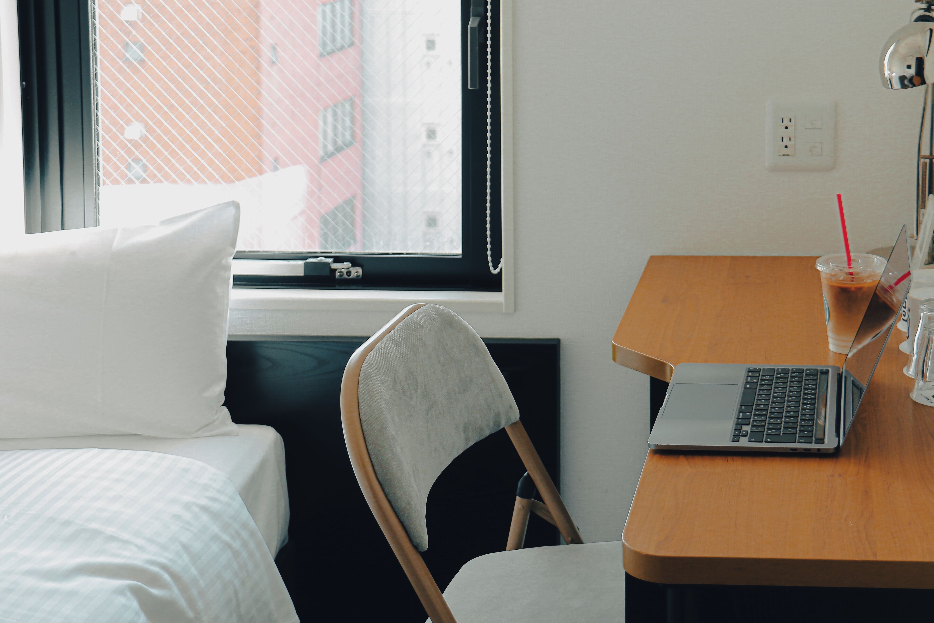 A hotel room with a bed, a desk with a laptop, a chair, a window with a view of buildings, and a bedside table with a drink and glasses.