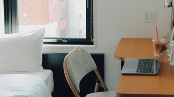 A hotel room with a bed, a desk with a laptop, a chair, a window with a view of buildings, and a bedside table with a drink and glasses.