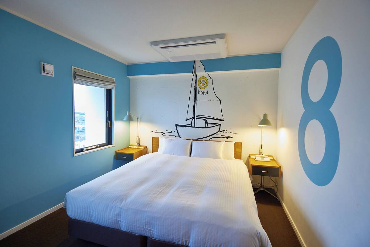Habitación de hotel con cama, ventana, aire acondicionado de pared, dos mesitas de noche con lámparas y un adorno de pared con un velero.