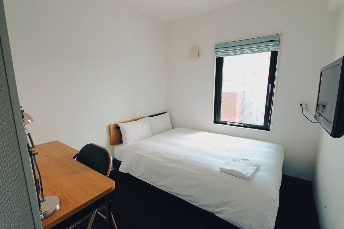 Una habitación pequeña y limpia con una cama individual, un escritorio y un ventanal con vista a edificios.
