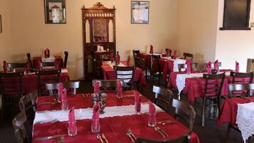 Uma sala de jantar com mesas preparadas para vários convidados, cada uma com toalhas vermelhas e toalhinhas de renda brancas.