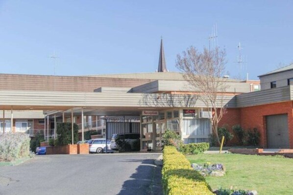 A motel with a covered parking area, a visible sign for 'Reception', and a church spire in the background. - Midcity Motor Lodge (Orange)