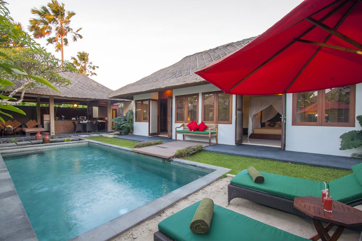 Uma área de piscina com espreguiçadeiras, um guarda-sol vermelho e um edifício com telhado de palha.
