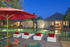 Zona de piscina con sillas de descanso y una mesa, rodeada por una terraza de madera y un paraguas rojo.
