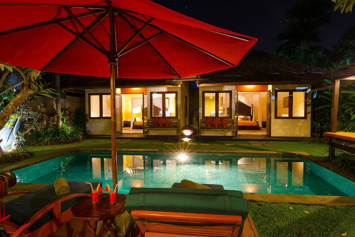Uma área na piscina com um guarda-sol vermelho, espreguiçadeiras e um prédio bem iluminado com várias divisões.