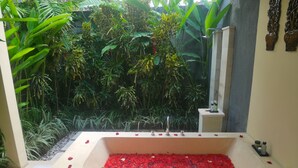 A rectangular bathtub filled with red flower petals, surrounded by lush greenery and a small white pedestal with toiletries. - Imani Villas (Kerobokan)