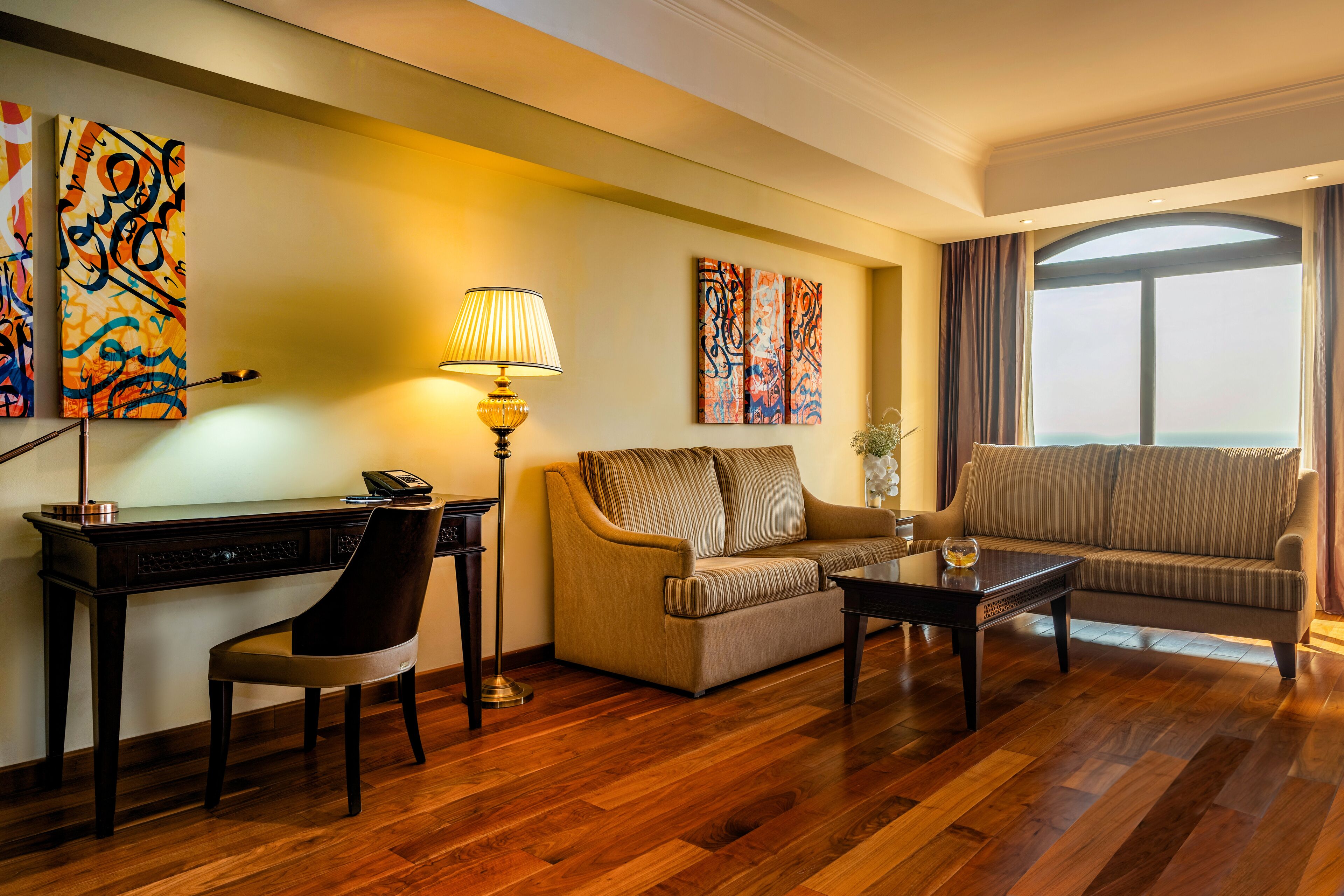 Habitación de hotel con piso de madera, un escritorio con silla, un sofá, una mesa de centro y dos obras de arte en la pared.