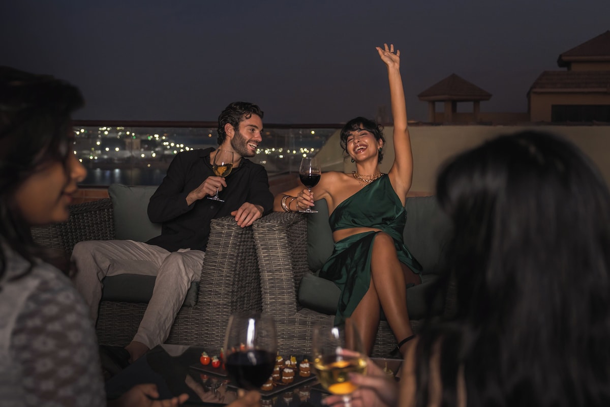 A rooftop terrace with wicker furniture, a table with wine glasses, and a city view at night.