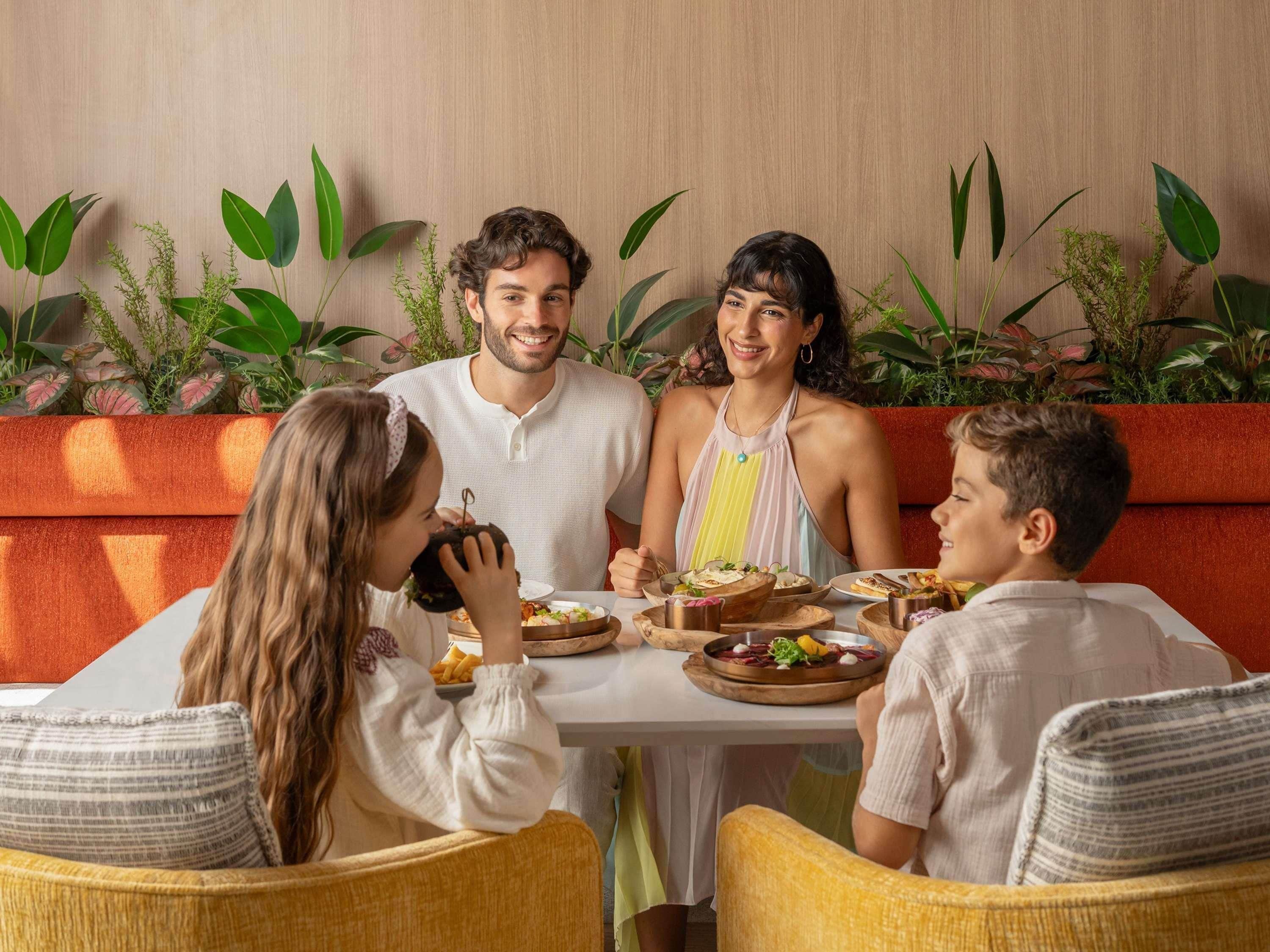 Eine Familie sitzt an einem Tisch und genießt eine Auswahl an Speisen, umgeben von viel Grün und einer Holzwand.